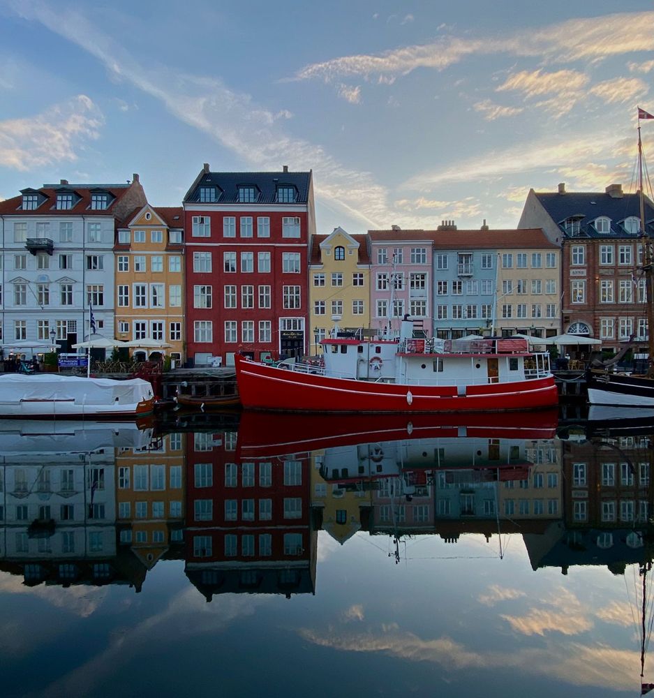 A view of Nyhavn, Copenhagen at sunrise, showing a row of colorful 17th- and 18th-century townhouses in shades of white, yellow, red, pink, and blue. A bright red and white boat is docked in the calm canal, perfectly mirrored in the still water below. The sky is soft blue with scattered clouds, and the reflections create a nearly symmetrical composition.