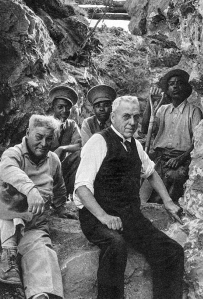 Black-and-white photograph of five men sitting and standing in a rocky excavation site. Two older men in the foreground wear light-colored shirts, one with a vest, while three men in the background, wearing work clothes and hats, are positioned among the rock walls, one holding a shovel. The man in a vest is pointing to a place in the rock where a fossil had been found.