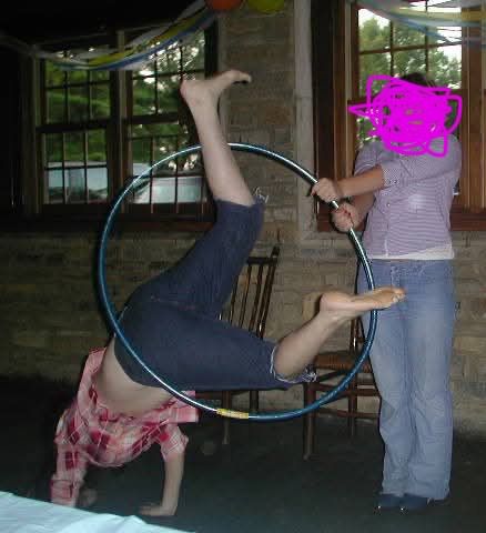 A photo me, wearing a pink and red plaid shirt, cutoff capri jeans and no shoes, attempting to leap through a hula hoop, held by a person with a marked out face.