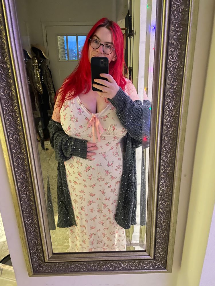 A mirror selfie of me wearing pale cream long dress that has a pink rose pattern on it. It has a pink silk ribbon with a bow on the neckline. Over it I am wearing a gray knitted cardigan with silver sequins. My hair is long and loose, and my head is tip to the side revealing more of my face, that I usually show. You can see my dark eyebrows, my greenish eyes with tortoise pattern, glasses, and my rosy cheeks and funny shaped nose, along with a bit of my thin lipped smile.