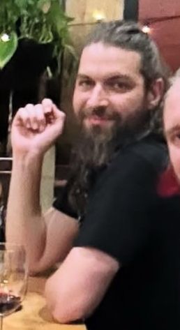 A blurry closeup of me, white guy with long hair and a beard, smiling vaguely, with elbows on the table and a distinct sass in the hand. 
