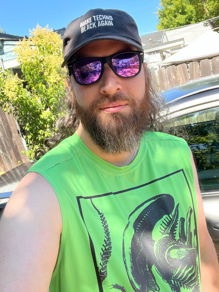 It’s me (white, beard, long hair) doing a selfie outside in a sleeveless green top with the Alien xenomorph. Purple and black sunglasses and a black cap with white letters reading “Make Techno Black Again”