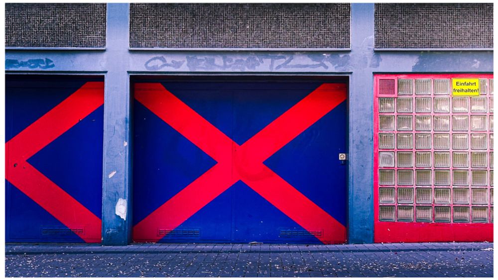 Querformat. Man sieht einen Teil einer Hauswand. Diese wird von schmalen Betonsäulen und -trägen in sechs Rechtecke unterteilt. Im Bildausschnitt ist nur das untere mittlere Rechteck vollständig zu sehen, die anderen sind abgeschnitten. In dem Rechteck befindet sich ein blaues Garagentor mit großem roten X. Links daneben ein identisches Tor, bei dem das x nur halb im Bild ist, rechts eine Glasbaustein-Wand mit rotem Rahmen. Darauf ein gelbes Schild mit der Aufschrift „Ausfahrt freihalten“.
Die oberen Rechtecke sind nur groben grauen Betobplatten gefüllt.
Auf der Wand sind schwache Spuren von entferntem Graffiti zu sehen.
Vor dem Haus ein schmaler Streifen aus Pflastersteinen