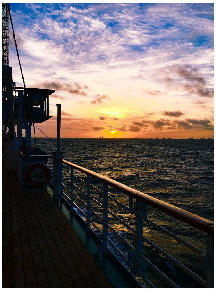 Hochformat. Das Bild eines Sonnenuntergangs über dem Meer. Man blickt die Reling eines Schiffes entlang. Diese führt von rechts unten in die linke Bildmitte. Die Planken liegen im Schatten und sind nur schemenhaft erlernbar. In der Bildmitte geht die Sonne unter. Der Himmel ist voller kleiner Wolken und hat einen satten Verlauf von orange zu blau (ganz oben im Bild) am Horizont sind kleine Silhouetten von anderen Schiffen zu sehen