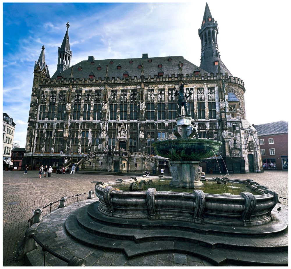 Querformat. Hauptelement des Bildes ist die Fassade des Aachener Rathauses. Davor der Brunnen mit der Figur Karls des Großen. Ein paar Menschen sind vereinzelt auf dem Marktplatz unterwegs. Links und rechts sind  ein paar andere Häuser zu sehen. Der Himmel ist blau mit Schleierwolken.