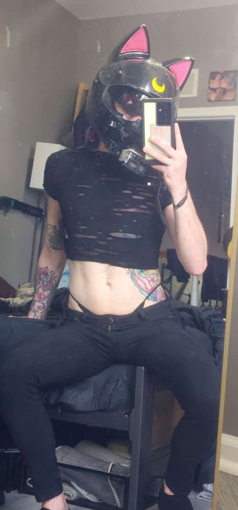 A cute femboy with a Luna x Celty motorcycle helmet poses in black tight jeans and a tight semi sheer crop top with visible piercings and straps. 