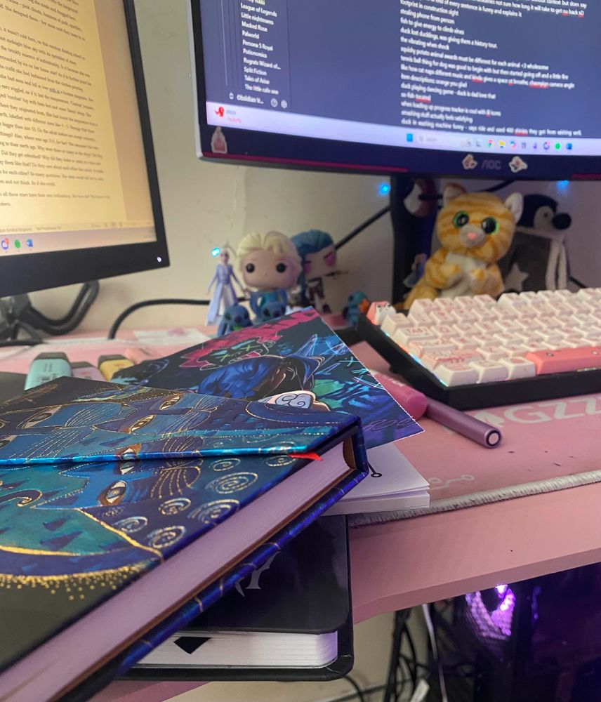 A picture showing three different notebooks, a night aesthetic cat one, a Jinx one and a Wednesday one. There are two screens, both with notes on. They are surrounded by highlighters, pens and figures of Elsa and Jinx. Also there sits a small orange striped cat teddy. 