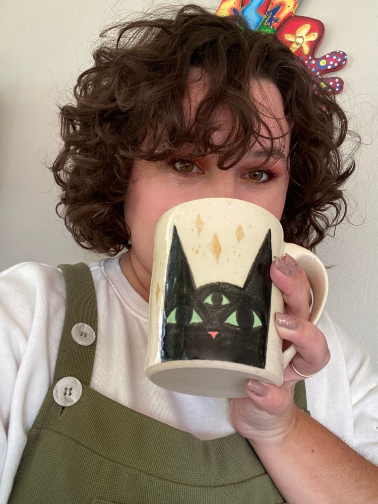 OP holding a mug with a black cat’s head on the front, complete with an open third eye on its forehead and yellow air sparkles. ✨ 