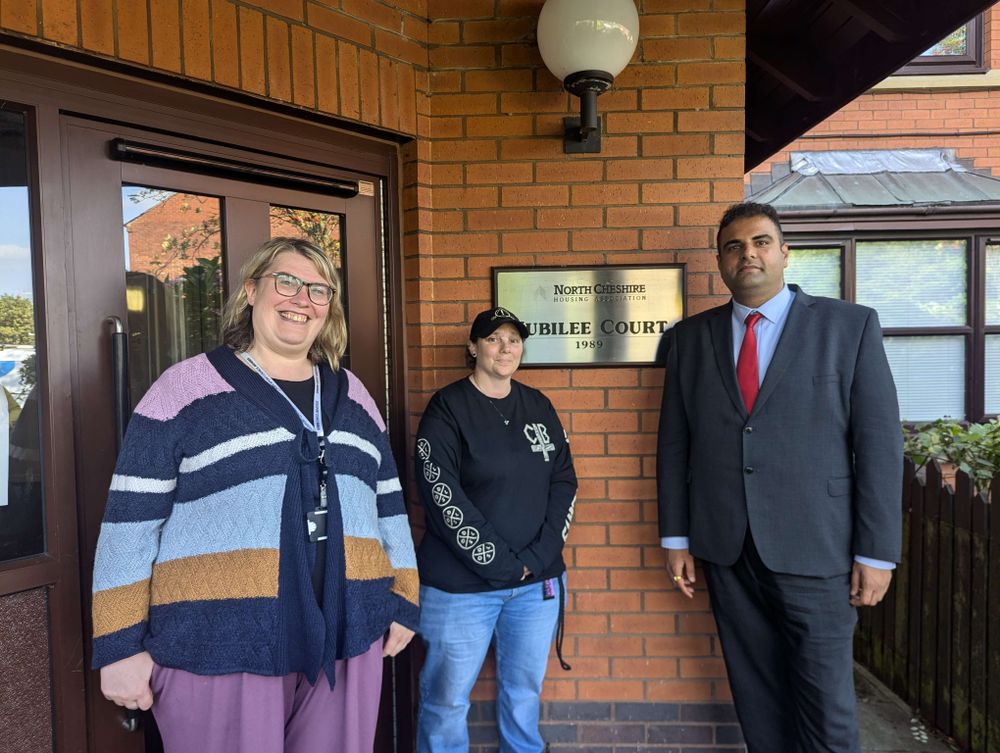 Three people stand smiling outside a building with a plaque that reads JUBILEE COURT