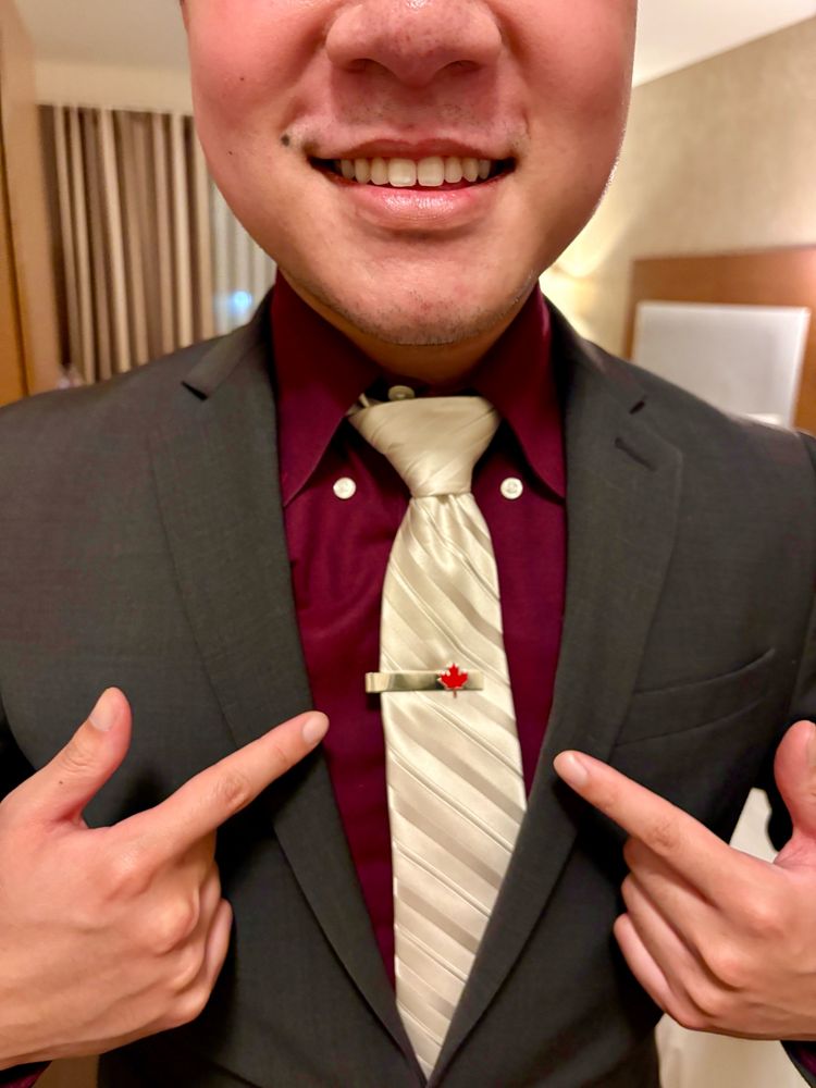 Viet is dressed in a dark gray suit with a red shirt, white tie, and a tie clip bearing a red maple leaf. 🇨🇦
