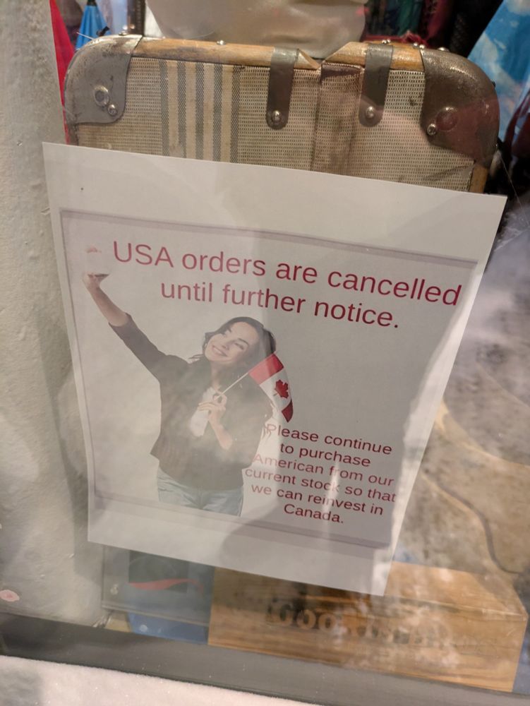A storefront notice that "USA orders are cancelled until further notice. Please continue to purchase American from our current stock so that we can reinvest in Canada." Alongside is an image of a woman taking a selfie while holding a Canadian flag.