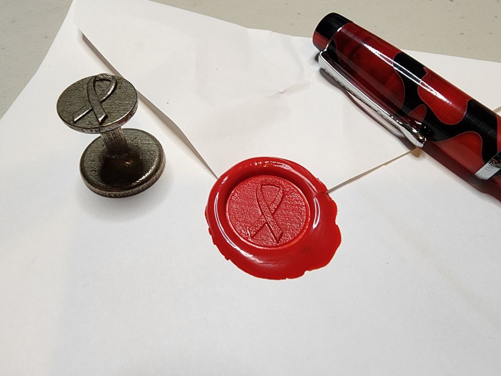 Photo of a white envelope with a red wax seal of the Awareness Ribbon, with a red & black pen & the stamp for the seal