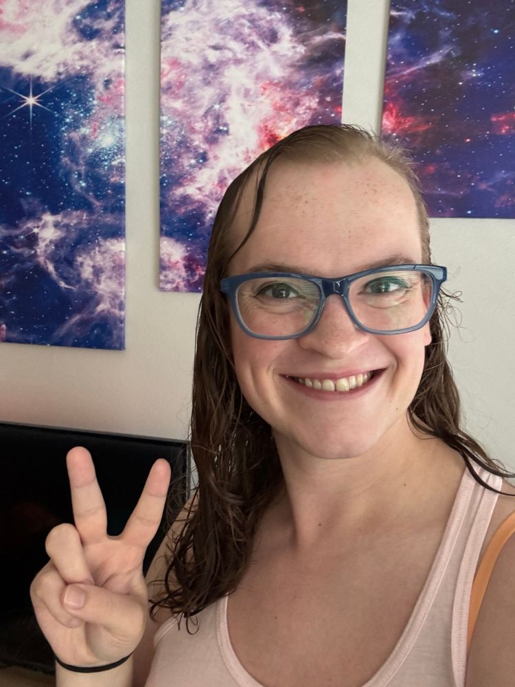 Taylor is wearing her pink tank top, with her long brown hair spilling over her right shoulder while the left side is tucked behind her shoulder. She has her classic peace sign and blue glasses on, and her background features some JWST imagery art