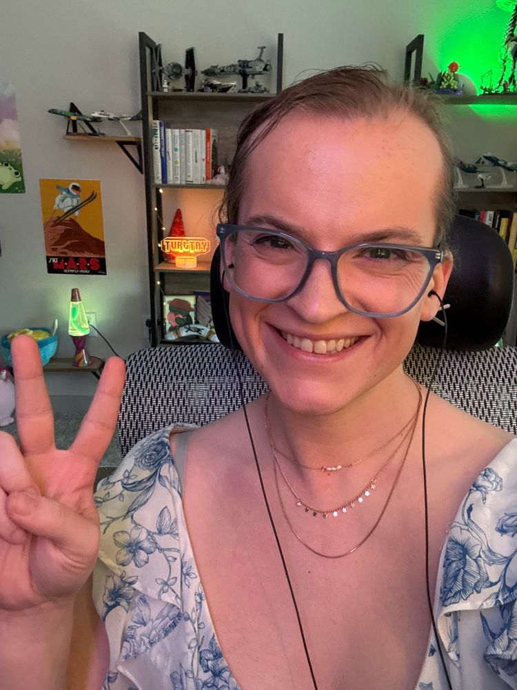 Tay has a big smile and is holding up a peace sign. She’s wearing a frilly dress with a white base and blue lacey trim. She also has a three-piece gold necklace. The daylight is diminishing, but there’s still enough to illuminate her bookshelf of cozy knick-knacks