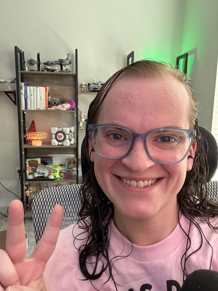 Tay is smiling and holding up a peace sign. She has her blue glasses on, which complement her curly, wet hair well. She’s wearing a pink T-shirt, and her background features a bookshelf of cute knick-knacks, including a 3D printed Companion Cube from Portal