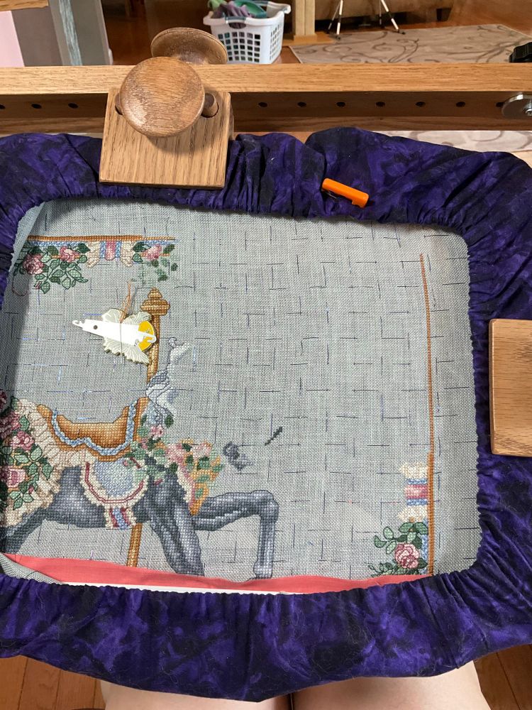 The same grey linen fabric. Now the back half and front legs of a black horse are present. The horse is wearing a golden saddle with a gold carousel pole. There are ivory bows in the center of the upper and right sides. The upper is back stitched and looks like a bow while the right is just a mush of pale color because it hasn’t been back stitched. 