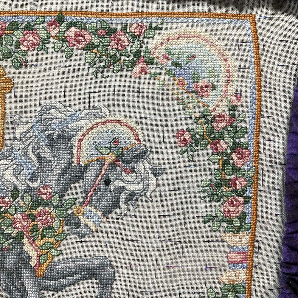 The upper right corner of a cross stitched carousel horse showing the front of the black horse and all the refugee roses from the Rose Garden. An ivory bow is in the mix on the right hand of the frame and a blue & ivory fan is in the upper right. Most of the work has been finished with back stitching, but the areas that aren’t are more suggestions of what is there than defined.