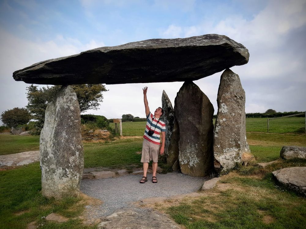 Photograph of the Neolithic Pentre Ifan dolmen in Wales. The remains of the chambered tomb consist of a large capstone balanced on the tips of three standing stones. A man stands underneath, one arm stretched upwards