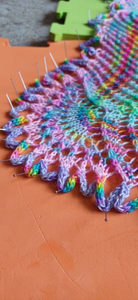 A close up of the very edge of a lace shawl, pinned to an orange blocking mat. A flower like lace cresent with i-cord loops is in the centre. The colours are pastel, with neon rainbow pops, the loops round the very edge are mauve with spaced rainbows.

Pattern was mashed together from several in order to use up all the yarn from an advent calendar. (Yarn dyer sells jkr merch so I won't list them)