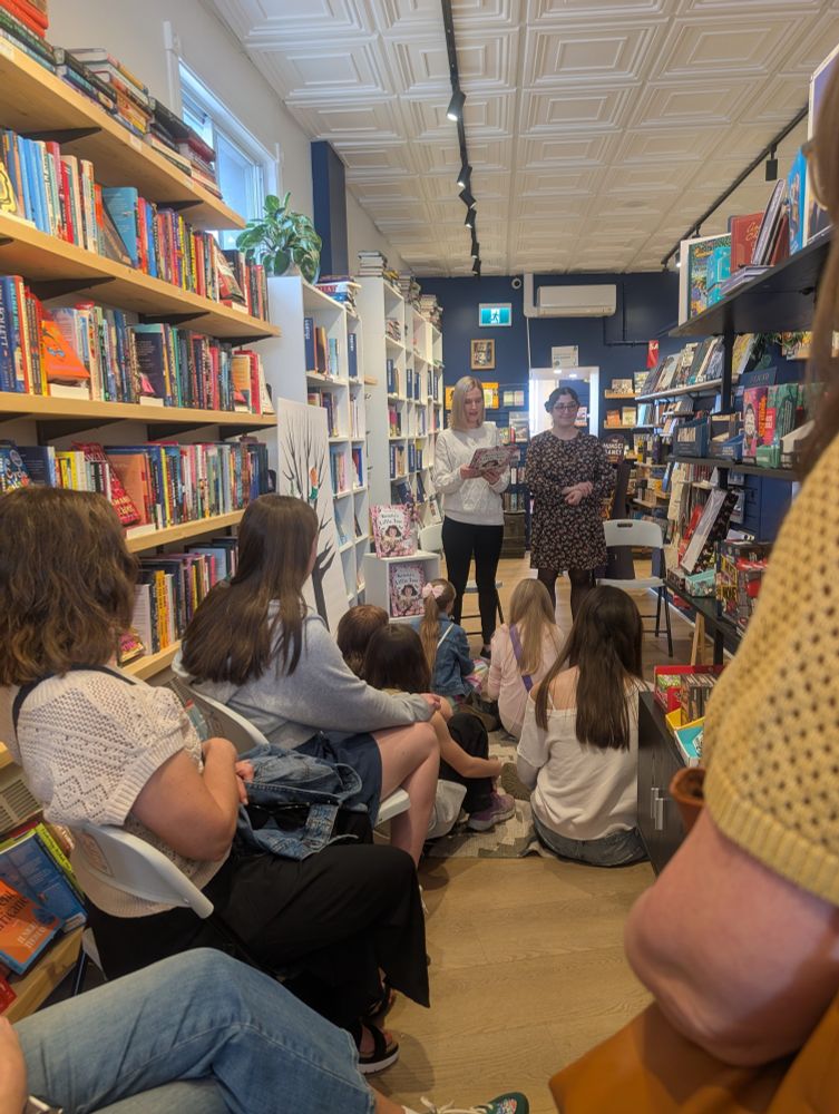 The author and illustrator welcome a group of seated children and adults to a book launch in a bookstore.