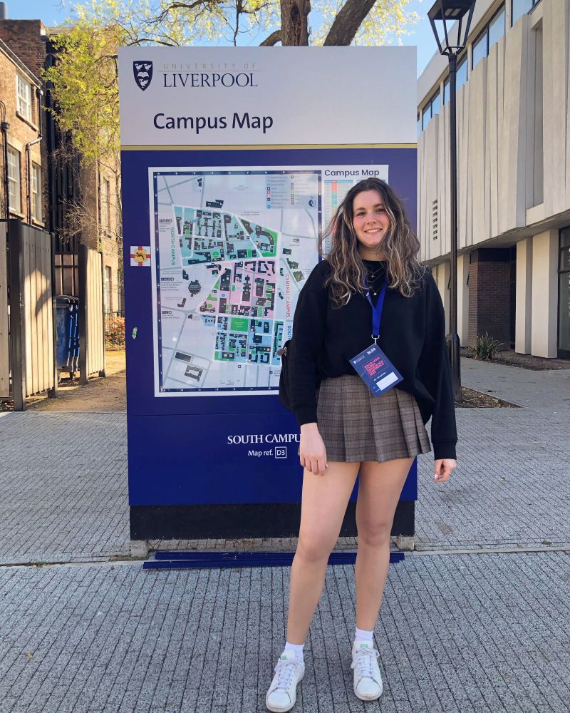 Person standing in front of a large “Campus Map” sign at the University of Liverpool, wearing a dark jumper, checked skirt, and conference lanyard. The setting is outdoors on a sunny day, with campus buildings and trees in the background.