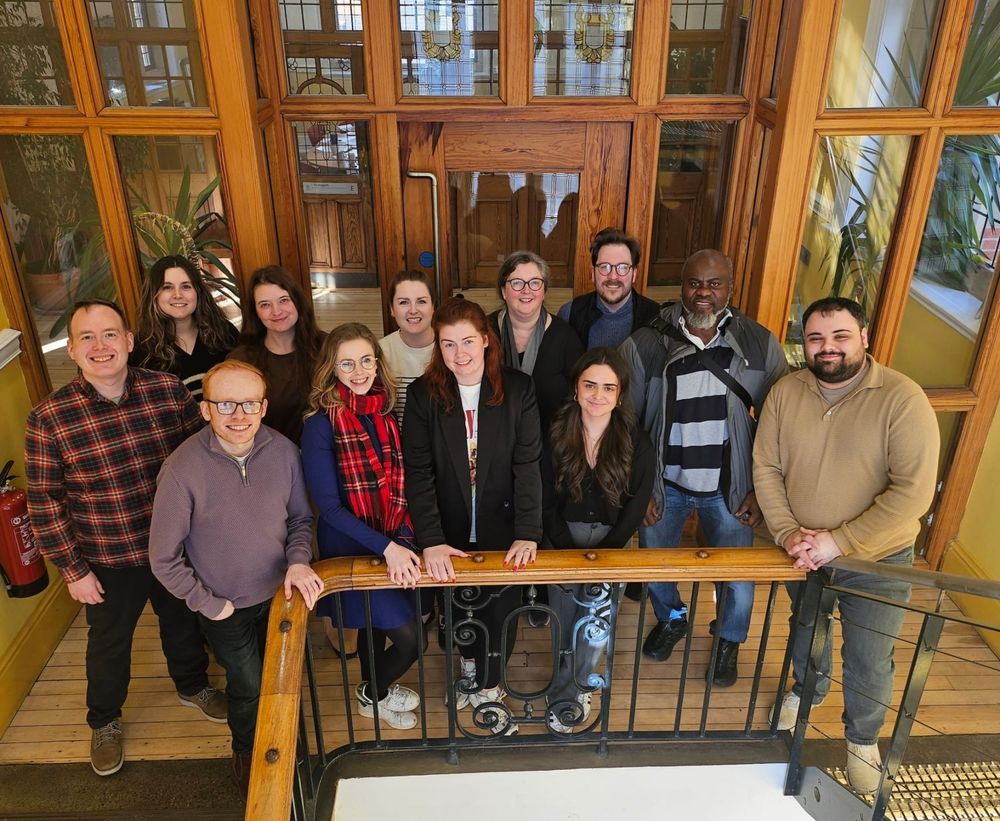 A group of 13 people stands smiling on a staircase inside a bright building with stained glass windows and wooden paneling. They are posing for a group photo, likely after a workshop or event. The setting has a warm, classic feel with natural light, tall plants, and vintage details. The group appears diverse in gender and age, dressed in smart-casual attire, and they look happy and engaged.