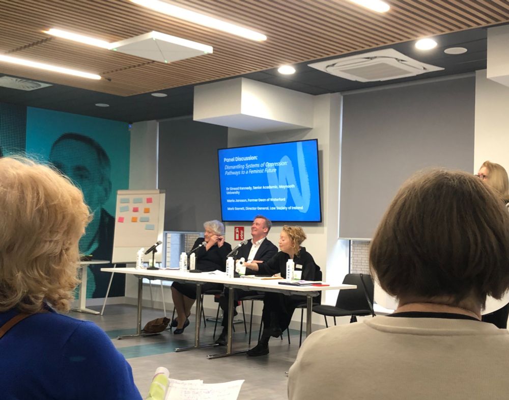 A panel discussion in a modern conference room with three speakers seated at a long table, engaging with the audience. The screen behind them displays the topic: 'Dismantling Systems of Oppression: Pathways to a Feminist Future.' Attendees, mostly women, are seated in rows, some taking notes. The room has contemporary wooden ceiling panels and bright lighting