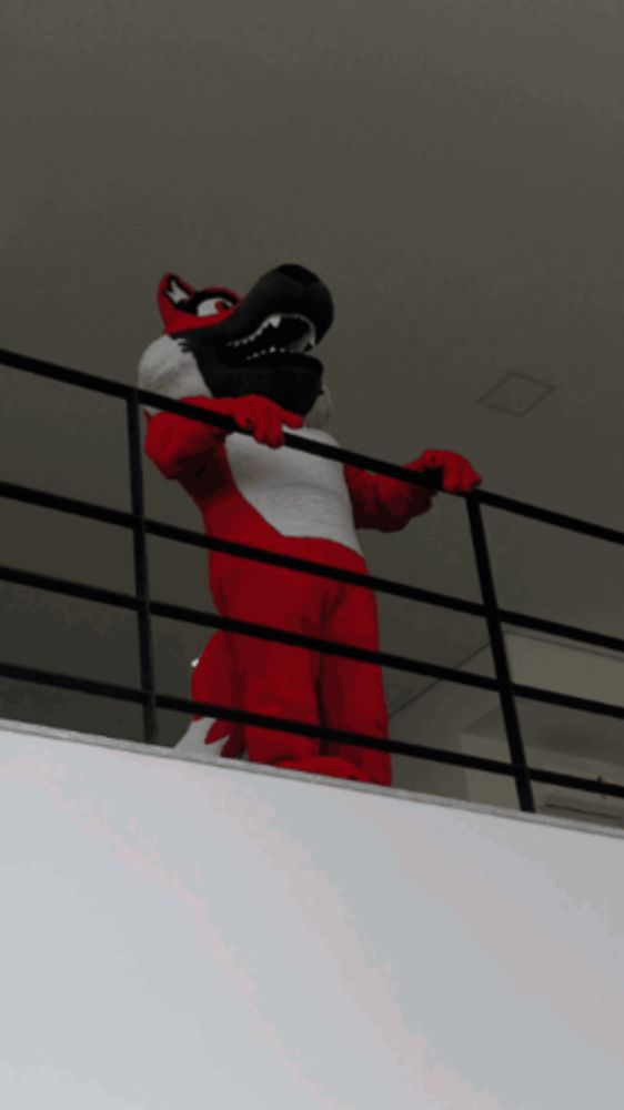 a red and white mascot is standing on a balcony with his arms outstretched