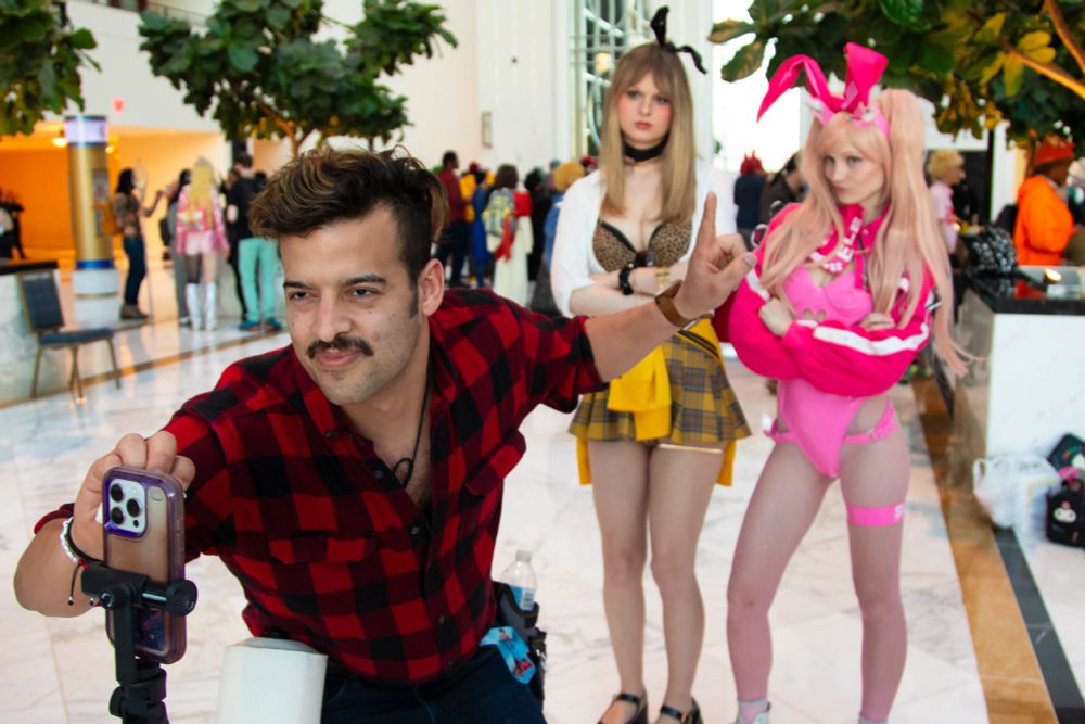 In the image, a man dressed in a red and black plaid shirt with rolled-up sleeves, cosplaying as the Brawny paper towel mascot, wiping a cellphone screen with a paper towel. He has a confident smirk, a thick mustache, and tousled dark brown hair. His left hand is raised, making a casual peace sign gesture, while his right hand holds the phone, which is mounted on a small tripod. A roll of paper towels is visible in front of him, reinforcing the humorous reference to the Brawny Man.

In the background, two cosplayers dressed as characters from the mobile game Goddess of Victory: Nikke stand with crossed arms, appearing mildly unimpressed or amused by the scene. The Rupee cosplayer on the left has long, straight blonde hair and wears a leopard-print bra, a white open blouse, and a yellow plaid skirt with matching accessories. A black headband with bunny ears is atop her head, and she sports black wristbands.

The Alice cosplayer on the right wears a bold pink bodysuit with a high-cut design, thigh-high stockings, a pink and white varsity-style jacket, and oversized pink bunny ears on her blonde pigtails. She has a playful yet skeptical expression, slightly pursing her lips.

The setting is a bright, spacious convention hall with a high ceiling, large windows, and marble flooring. Various convention attendees in colorful outfits and costumes can be seen in the background, engaging in conversations and posing for pictures. The area is decorated with indoor plants, and natural light streams in, enhancing the vibrant atmosphere. The interaction between the three main subjects adds a comedic dynamic to the scene, as the Brawny Man cosplayer theatrically cleans the phone while the two Nikke cosplayers react with annoyance.