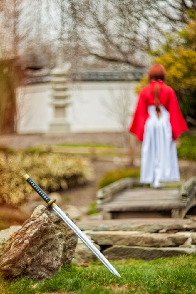 A photograph with a cinematic and atmospheric quality features a scene in a tranquil garden setting. In the foreground, a traditional Japanese katana sword with a sharp, gleaming silver blade and a black handle with gold accents is embedded into the grass, leaning against a textured stone. The sword's placement and intricate detailing make it a focal point of the image.

In the background, a figure dressed in traditional Japanese attire—a red haori (kimono jacket) and flowing white hakama (wide-legged pants)—stands with their back to the viewer. The character's long red hair cascades down their back, emphasizing the serene yet enigmatic mood. The figure is positioned on a small wooden bridge, with the garden's natural elements—mossy rocks, wooden planks, and soft greenery—surrounding them.

The shallow depth of field blurs the background slightly, creating a sense of separation between the sword in sharp focus and the figure, adding mystery and narrative intrigue. The muted, earthy tones of the garden contrast with the vibrant red of the figure's attire, evoking a timeless, contemplative, and dramatic atmosphere. The overall composition, with its blurred background, vivid focus on the katana, and natural surroundings, gives the image an almost poetic and cinematic quality.