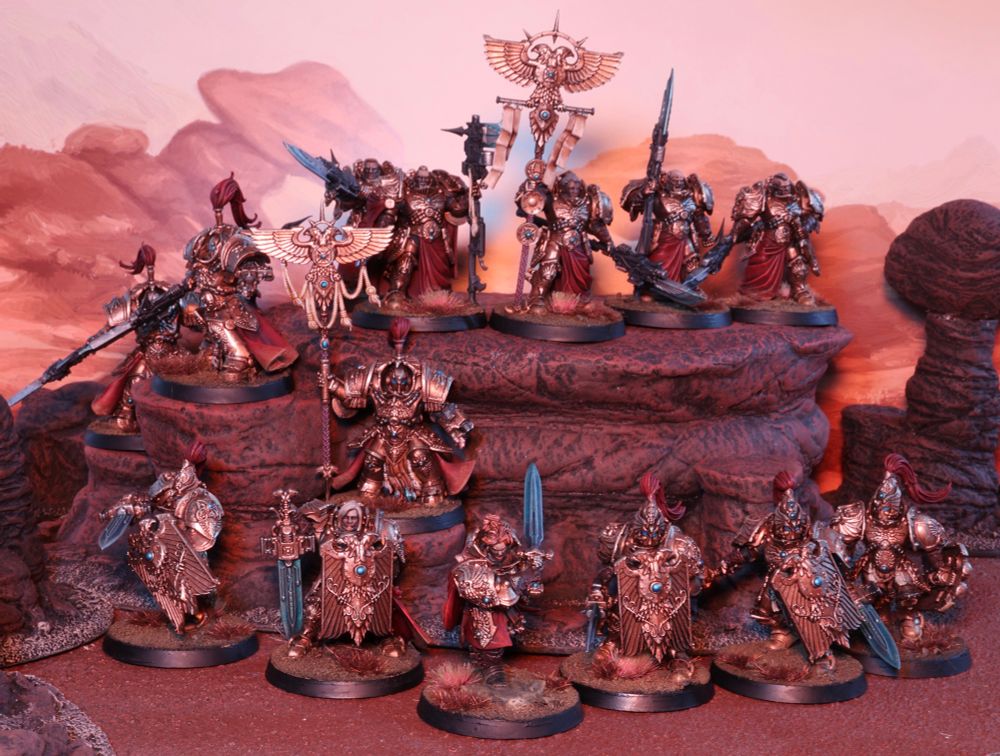 A collection of Adeptus Custodes miniature figures, by Games Workshop, is arranged on simulated rocky terrain. The figures, resembling heavily armoured warriors, display gold and red colour schemes, adorned with ornate designs and blue gemstones. Some wield weapons like swords and spears, while others hold banners. The background features a painted desert landscape in warm tones. This version is cast in a moody orange light, suggesting an alien world or irradiated landscape
