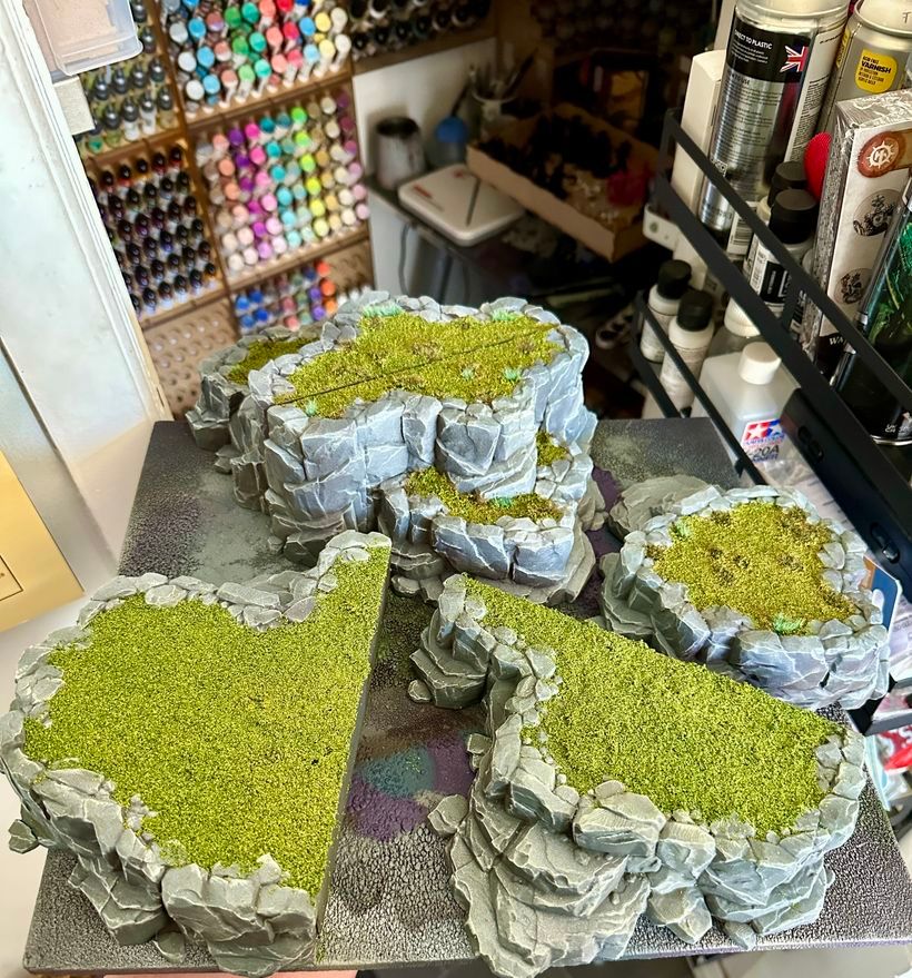 A collection of model rock formations is displayed on a grey base, featuring intricately textured grey rocks with vibrant green grass on top. The formations vary in shape and size and are displayed amid an artist's workspace, visible with numerous colourful paint bottles in the background.