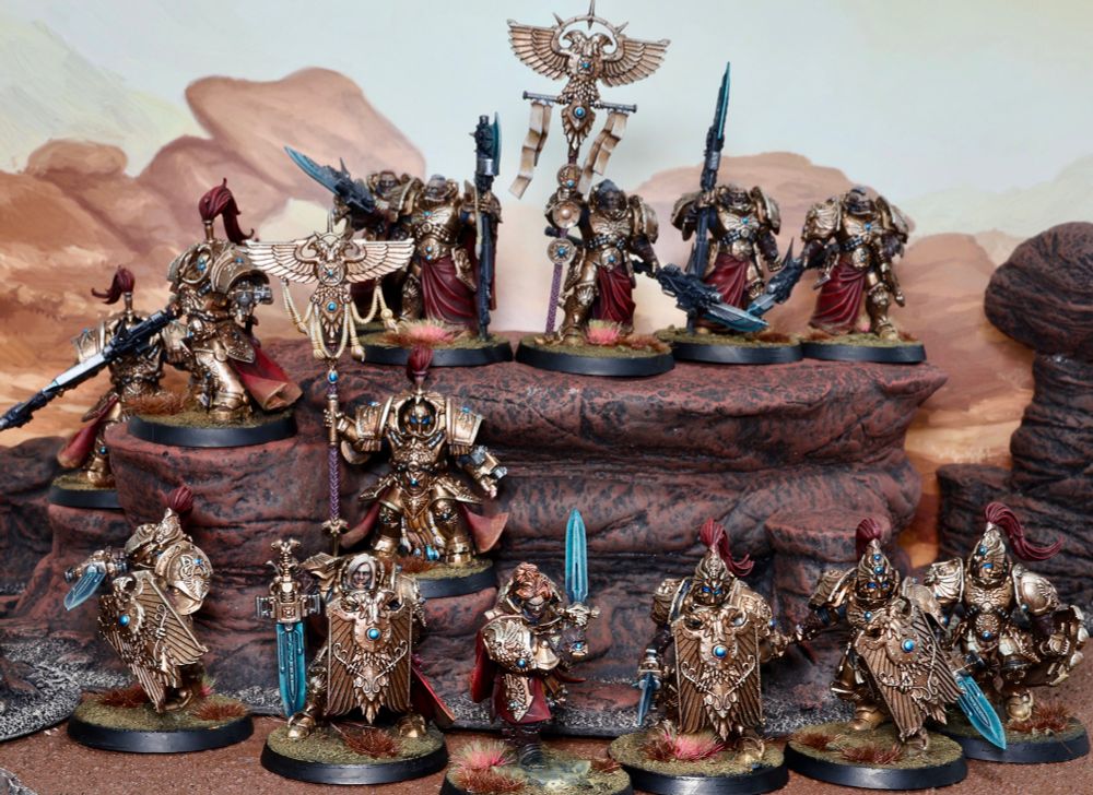 A collection of Adeptus Custodes miniature figures, by Games Workshop, is arranged on simulated rocky terrain. The figures, resembling heavily armoured warriors, display gold and red colour schemes, adorned with ornate designs and blue gemstones. Some wield weapons like swords and spears, while others hold banners. The background features a painted desert landscape in warm tones.