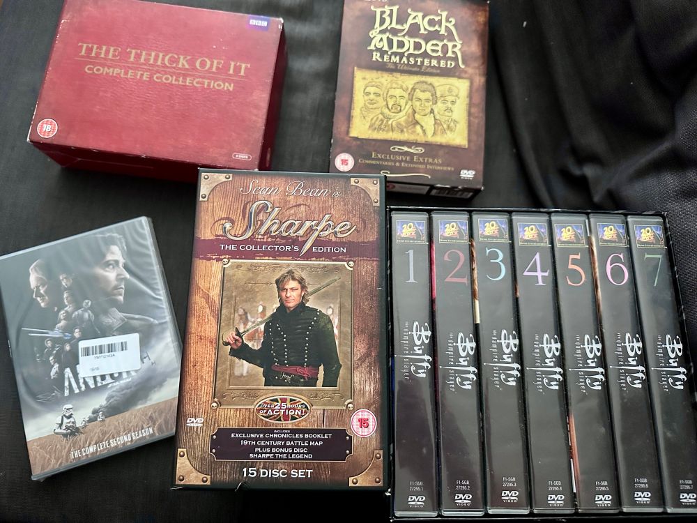 DVD box sets for: The Thick of It, Blackadder, Buffy The Vampire Slayer, Sharpe, and Andor season 2, arranged on a dark grey sofa