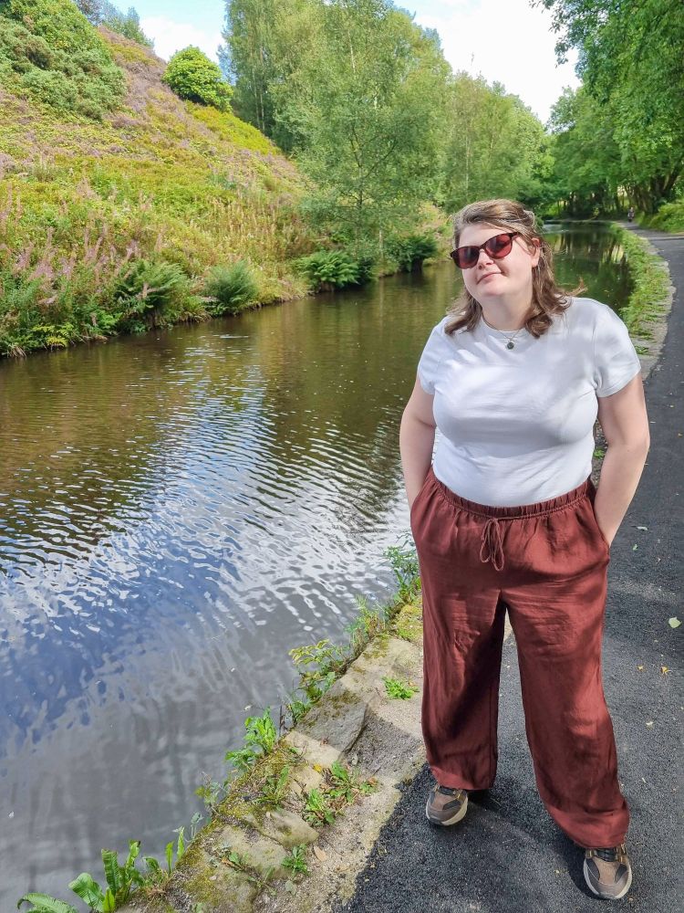 me stood next to a canal. im wearing a white t shirt, brown trousers, brown walking shoes, and sunglasses. on the other side of the canal there's a bank sloping up sharply, covered in foliage in various shades of green and purple.