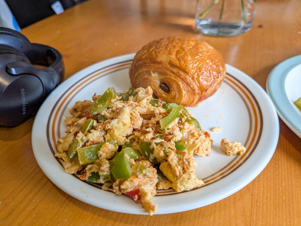 a plate of slightly overdone scrambled eggs with chunks of green pepper, tomato and tomatoes, with a gleaming pain au chocolate on the side.