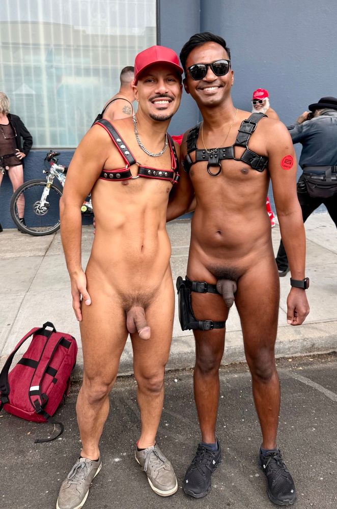 Two nude men wearing harnesses smile and stand together on a sunny street in San Francisco. Their posture is relaxed and open, suggesting an easy new friendship formed in a moment of shared freedom. The city buzzes in the background, adding to the sense of confidence, community, and celebration
