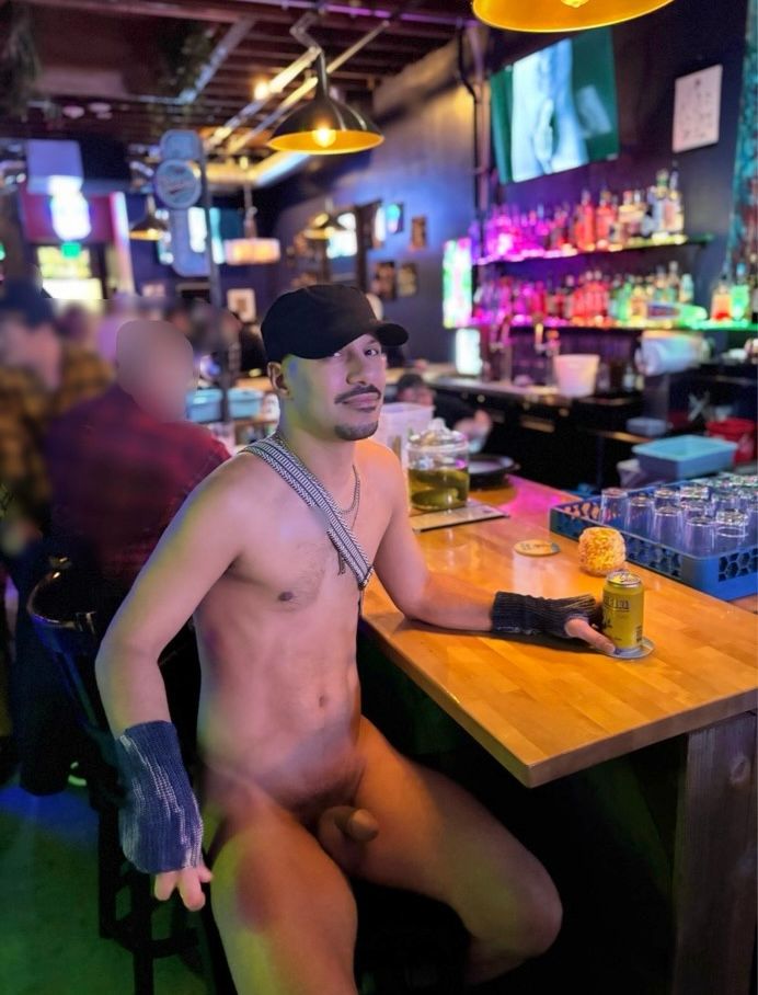 A naked man stands confidently at a bar, wearing nothing but a black cap and the gray knitted gloves he made himself. He holds a drink casually in one hand, showing off the gloves with pride, while the rest of his body is bare. Around him, other bar patrons mingle and chat, some noticing him, others carrying on with their evening, creating a playful contrast between his bold exhibition and the everyday social setting.