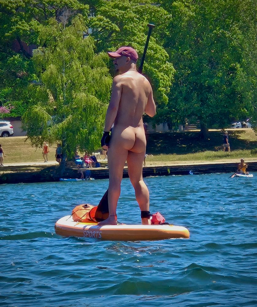 A naked man stands confidently on an orange paddleboard in the middle of a calm lake, his back to the camera and his bare butt facing the sunlit park shoreline. He wears a red cap and a black glove, holding a paddle as he looks ahead. Behind him, people relax on the grass beneath tall trees on a bright summer day.