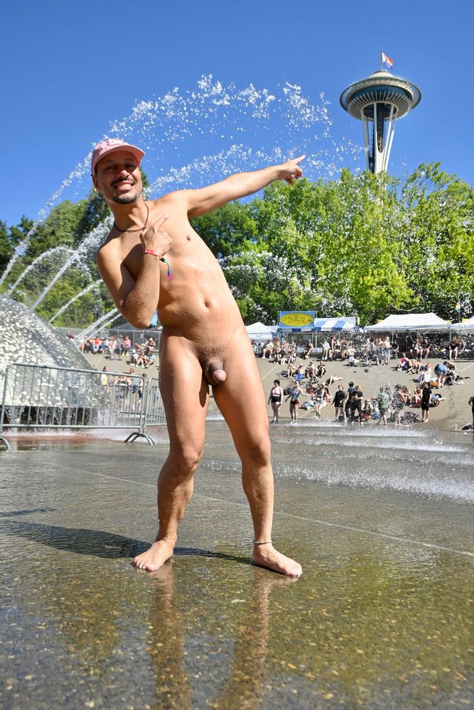 A smiling nude person wearing a pink cap stands barefoot in front of a large fountain at Seattle Center, pointing toward the Space Needle in the background. There are people sitting and playing around the fountain on a sunny day.