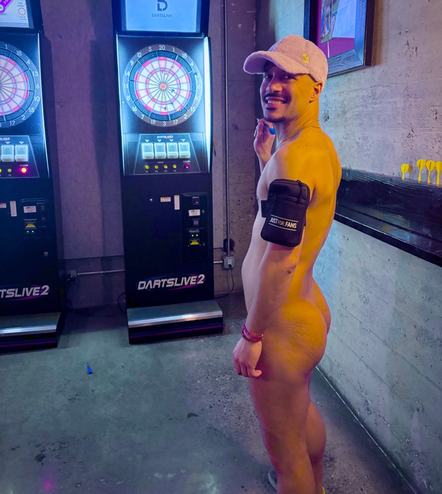 A nude man wearing only a pink baseball cap and sneakers stands in front of an electronic dartboard machine in a dimly lit bar. He smiles over his shoulder at the camera, holding a dart in his right hand, with his bare backside visible under the purple-blue lighting.