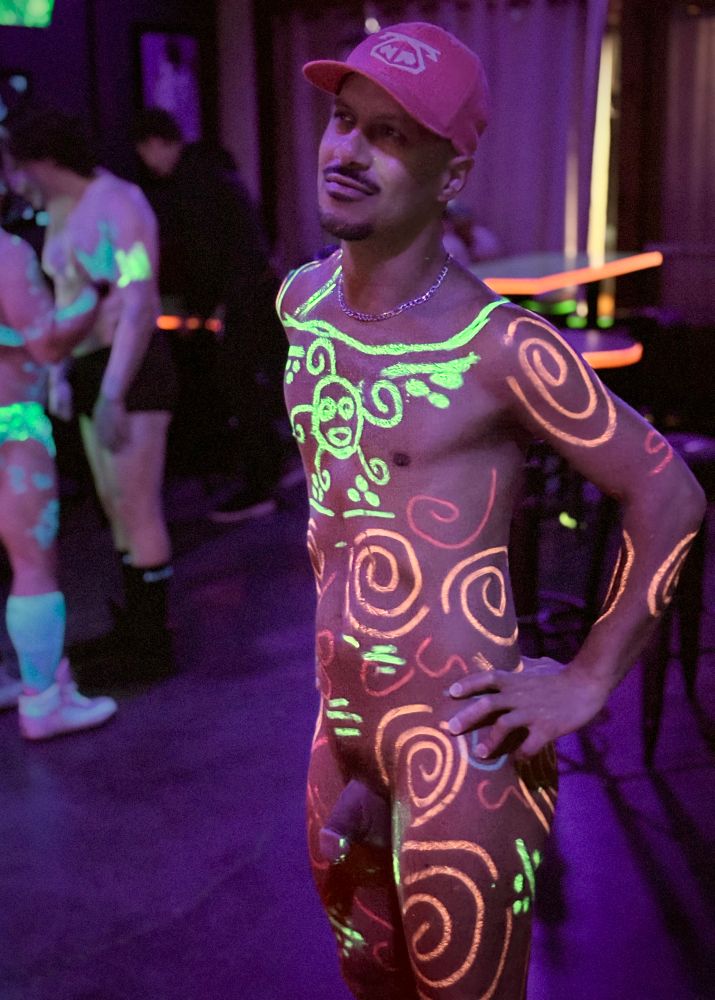 A nude man stands confidently in a dimly lit club illuminated by blacklight. His body is decorated with glowing Taíno-inspired body paint in swirling orange and green patterns, including a stylized sun face on his chest, while his circumcised penis remains unpainted. He wears a red baseball cap and a silver chain necklace, one hand resting on his hip. The neon paint glows vividly under ultraviolet light, with other minimally clothed people visible in the background.