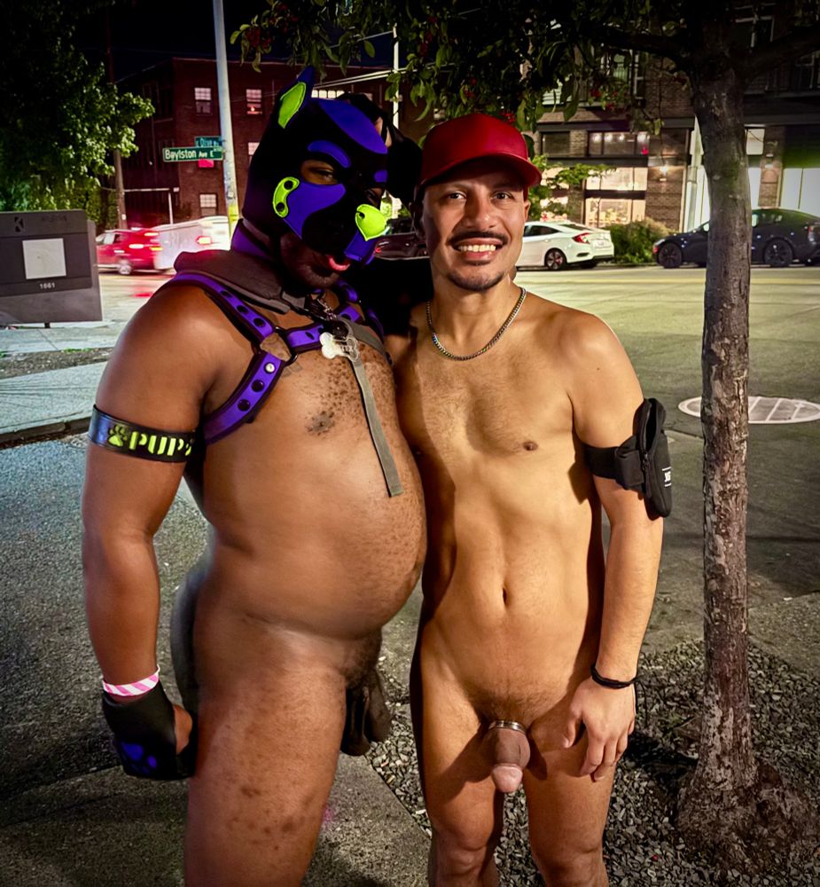 Two men pose together naked on a city sidewalk at night under a streetlight. The man on the left wears a black and purple pup hood, a matching harness, and black gloves, with one arm around the other man. The Latino man on the right smiles at the camera, wearing a red cap, a silver chain, an armband, and a cock ring. Cars and buildings with lit windows are visible in the background.