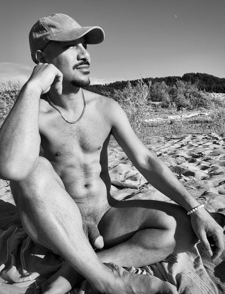 A black and white photo of a nude man sitting cross-legged on a sandy beach, wearing only a baseball cap, a chain necklace, and a beaded bracelet. He gazes into the distance with his hand resting on his face. Behind him are shrubs, dunes, and a forested hillside under a clear sky with the moon faintly visible.