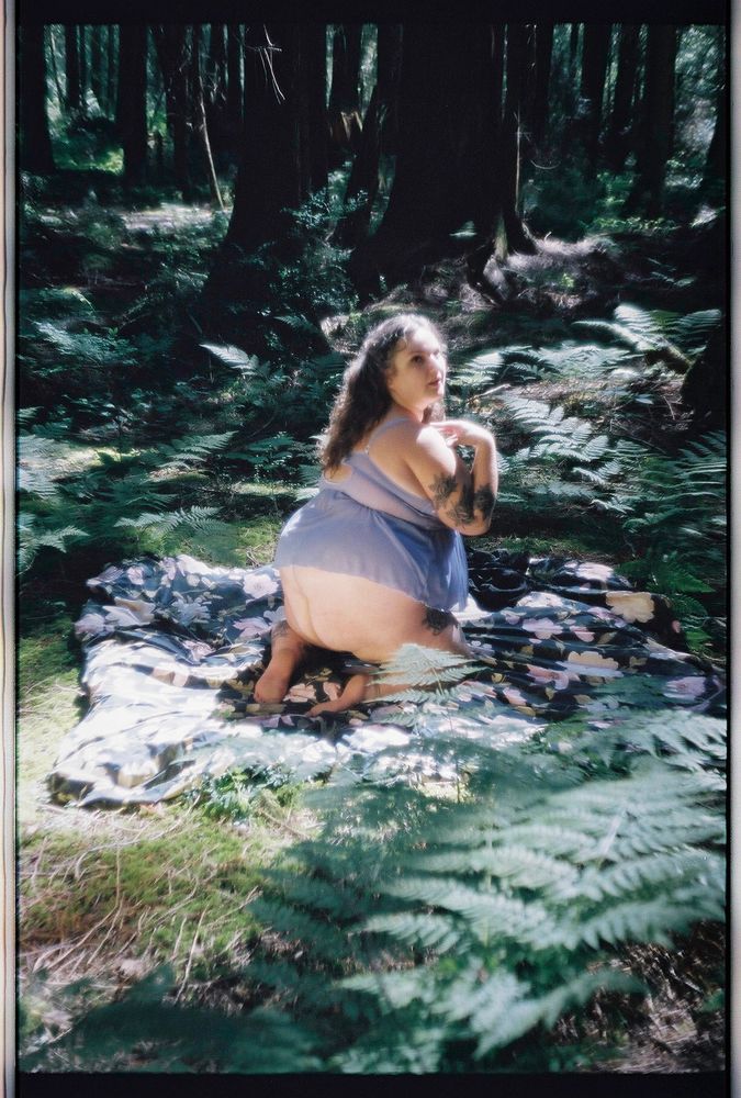 A photo of Sofia in a dense forest lush with dark green ferns. She is on her knees facing away from the camera and kneeling on a black floral quilt, but looking back over her shoulder at the camera. Soft golden light falls over her in her soft purple lingerie