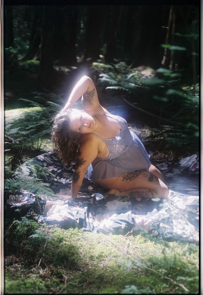 Sofia pictured on her knees in a backbend on the forest floor on a black and floral comforter. She wears a lilac sheer dress and all her long brown curly hair falls to one side of her face. The light is golden and dappled on her soft radiant body 