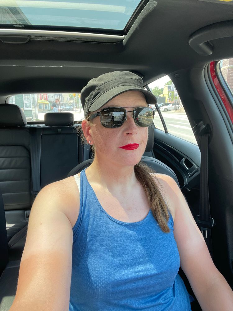 Cailín is sitting in her car. She’s wearing a black military style cap, black sunglasses, red lipstick, and a blue grey tank top 