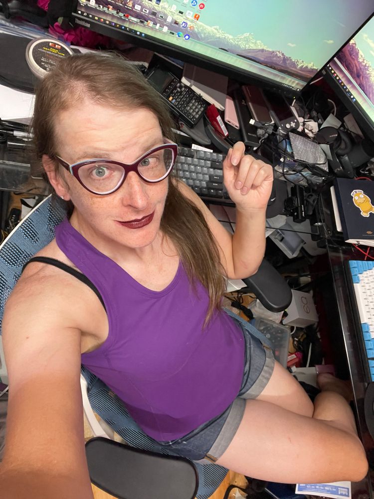 Cailín is sitting at her desk. She’s wearing purple cats eye glasses, burgundy lipstick, a purple tank top, and blue denim shorts with rolled cuffs 