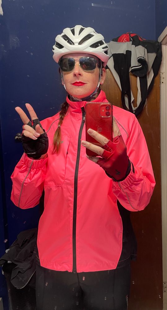 A mirror selfie. I’m wearing a white road cycling helmet, black sunglasses, red lipstick, a bright pink rain jacket, red and black cycling gloves, and black cycling shorts