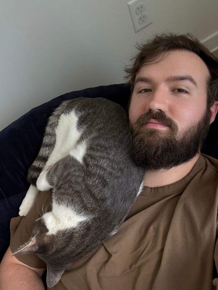 A severely sleep deprived (and slightly swollen) Andrew with an unkempt beard laying on a beanbag chair with a sleepy cat on his chest.